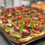 Tray of mini beef sliders topped with greens and sliced strawberries, ideal for gourmet catering, wedding platters, or corporate events